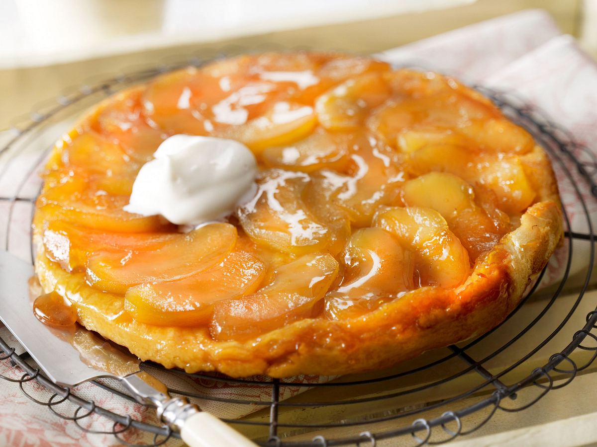 Tarte Tatin mit karamellisierten Apfelscheiben und einem Klecks Sahne liegt auf einem Kuchengitter, daneben ein Messer.