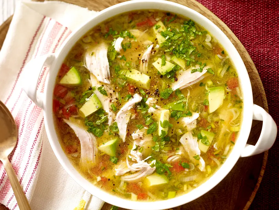 Texmex-Hühnersuppe Hühnersuppe mit zarten Hähnchenstreifen, Avocadowürfeln, Tomaten und frischen Kräutern, serviert in einer weißen Schale auf einem runden Holzbrett mit goldener Löffel und ein paar Tortillachips als Beilage.