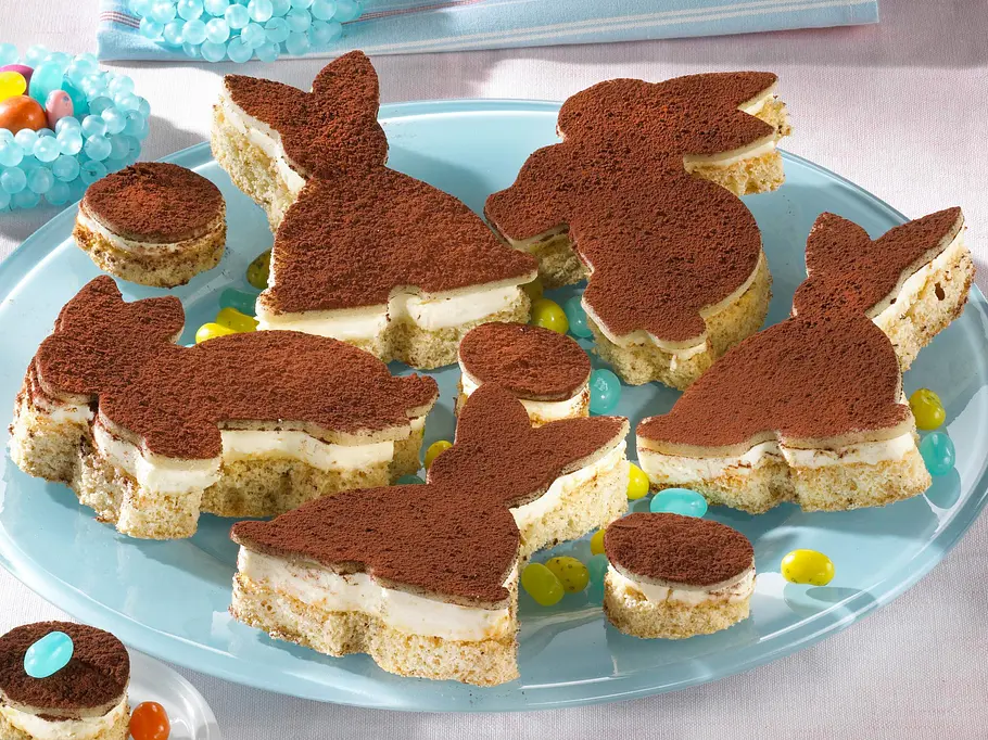 5 Tirmaisu-Hasen und 3 runde Tiramisu-Häppchen auf einem blauen Teller angerichtet mit gelben und blauen Bonbons.