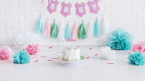 Eine Girlande mit Baby-Schriftzug auf weißer Wand, davor eine Torte und bunte Deko - Foto: iStock/Tatyana Maximova