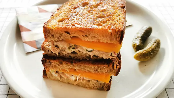 2 goldbraun gebacken Tuna-Melt-Sandwiches übereinander liegend auf einem hellen Teller, am Rand zwei Gewürzgurken - Foto: LECKER @ Bauer Media Group / Birte Wloka