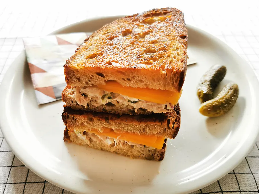 2 goldbraun gebacken Tuna-Melt-Sandwiches übereinander liegend auf einem hellen Teller, am Rand zwei Gewürzgurken