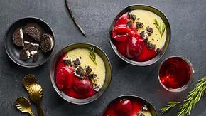 Drei Schalen mit cremigem Vanillepudding, getoppt mit roten Pflaumenhälften, zerbröselten dunklen Cookies und einem Zweig Rosmarin, daneben ein Schälchen mit weiteren Cookies und goldene Löffel auf dunkler Unterlage. - Foto: LECKER @ Bauer Media Group