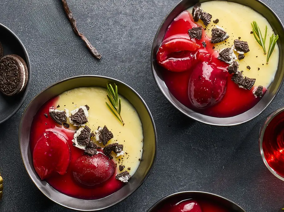 Drei Schalen mit cremigem Vanillepudding, getoppt mit roten Pflaumenhälften, zerbröselten dunklen Cookies und einem Zweig Rosmarin, daneben ein Schälchen mit weiteren Cookies und goldene Löffel auf dunkler Unterlage.