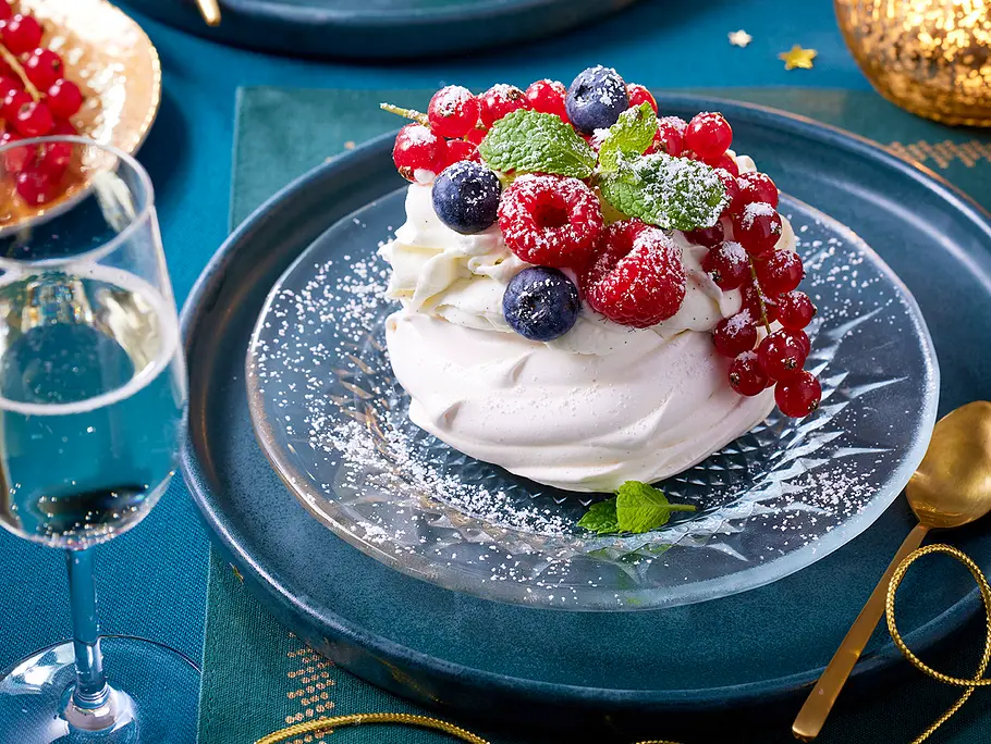 Verschneite Pavlova Rezept Auf einem kleinen Dessertteller aus Glas liegt eine Pavlova mit Beerentopping, leicht bestäubt mit Puderzucker
