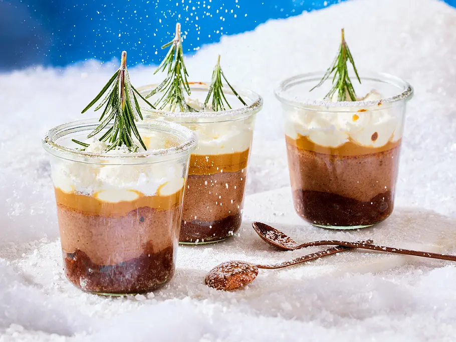 Drei kleine Weckgläser gefüllt mit Schichten aus Cheesecake und Sahnehaube mit kleinem Tannenzwei als Deko, rundherum Deko-Schnee
