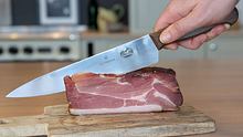 Großes Küchenmesser schneidet durch Schinken auf einem Holzbrett. - Foto: LECKER.de/Emily Beckmann