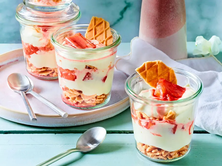Drei Gläser mit Schoko-Creme und Rhabarberkompott, garniert mit Waffeln, stehen auf einem Teller mit Löffeln auf einem Holztisch.
