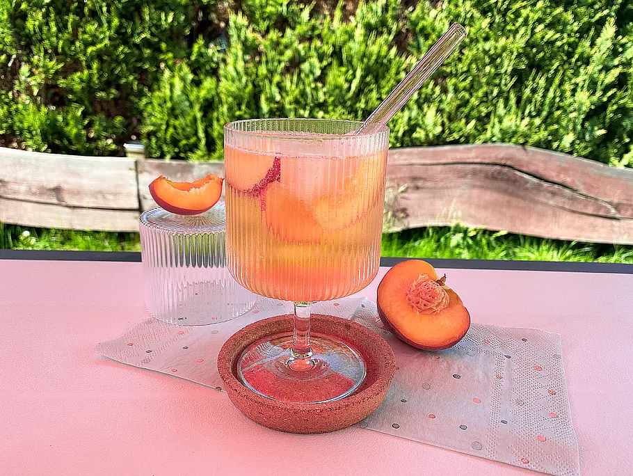 White Peach Spritz Rezept Ein geriffeltes Weinglas mit Peach Perfect Spritz steht auf einem weißen Tisch, im Hintergrund ist ein Gartenzaun und eine grüne Hecke zu erkennen