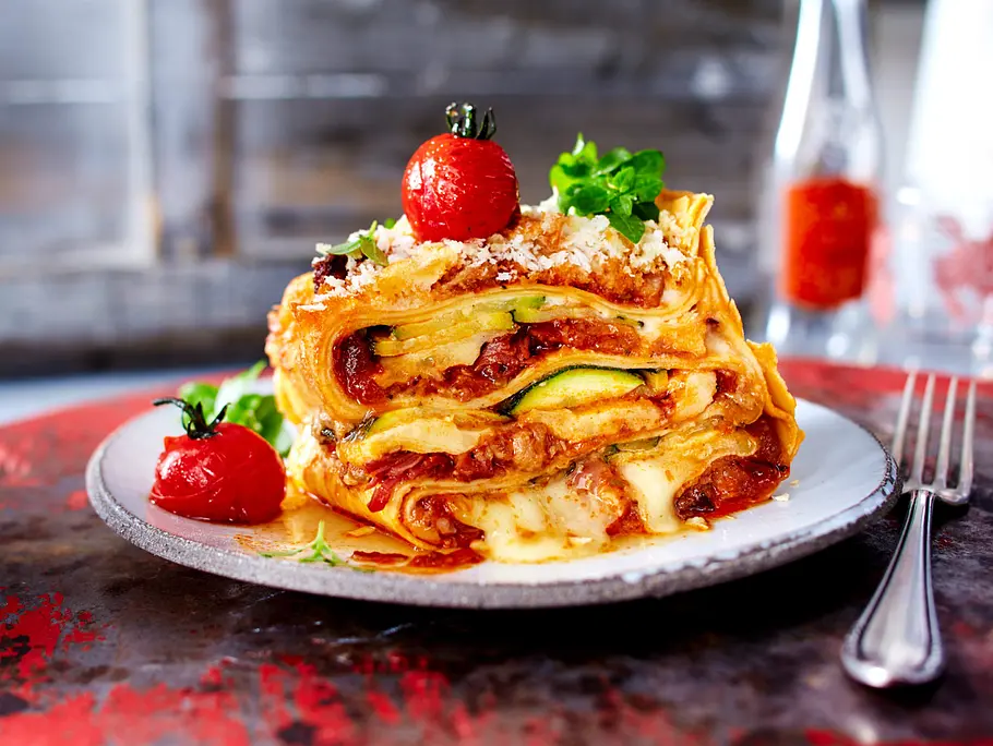 Ein Stück Lasagne mit Zucchini, Tomatensauce und Käse liegt mit kleinen Tomaten und Kräutern auf einem Teller.