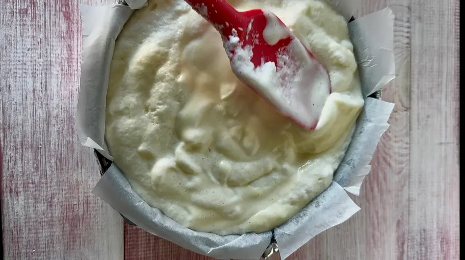 Wölkchenkuchen Schritt 2 Ein roter Teigschaber liegt auf hellem, schaumigem Teig in einer runden, mit Backpapier ausgelegten Form auf einem Holztisch.
