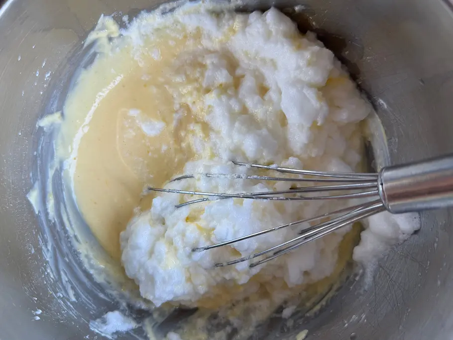 Wölkchenkuchen Schritt 1 Ein Schneebesen liegt in einer Metallschüssel mit gelblichem Teig und noch nicht vollständig untergerührtem Eischnee.