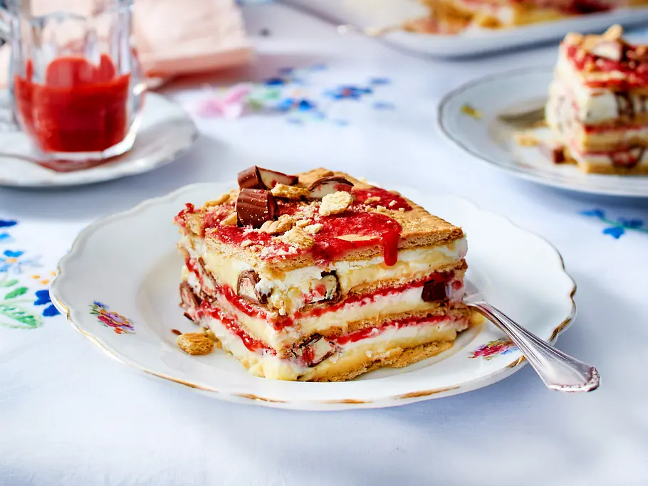 Ein Kuchenstück mit vielen Schichten aus Keks, Erdbeersoße und Pudding befindet sich auf einem klassischen weißen Kuchenteller mit Goldrand.