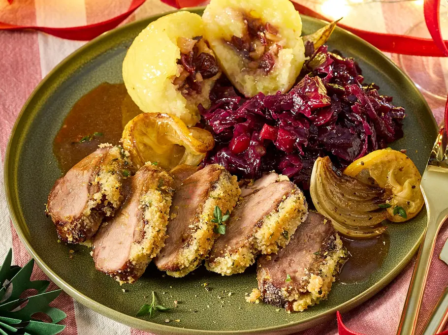 In Scheiben geschnittene Filets mit knuspriger Thymian-Kräuterkruste auf einem grünen Teller, daneben zwei gefüllte Klöße, Rotkohl und Bratensauce, serviert auf einer gestreiften Stoffdecke.