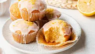 Vier Zitronenmuffins befinden sich auf einen hellen Teller. Jeder ist mit einer dünnen Schicht Zuckerguss und Zitronenraspel dekoriert. Der Muffin im Vordergrund ist angebissen. - Foto: ShowHeroes für LECKER @ Bauer Media Group