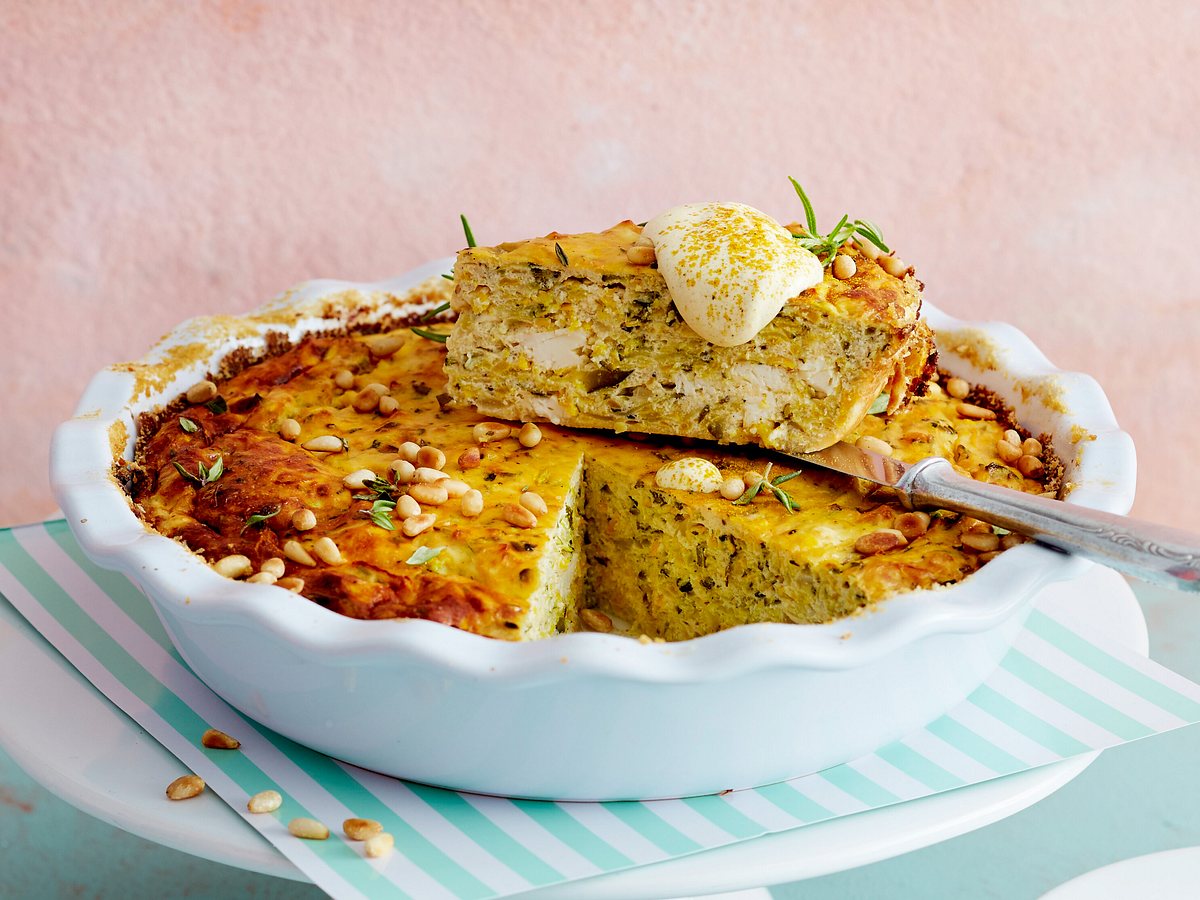 Ein Stück Zucchini-Feta-Kuchen mit Pinienkernen und Kräutern liegt auf einer Kuchenform, darauf ein Klecks Creme.