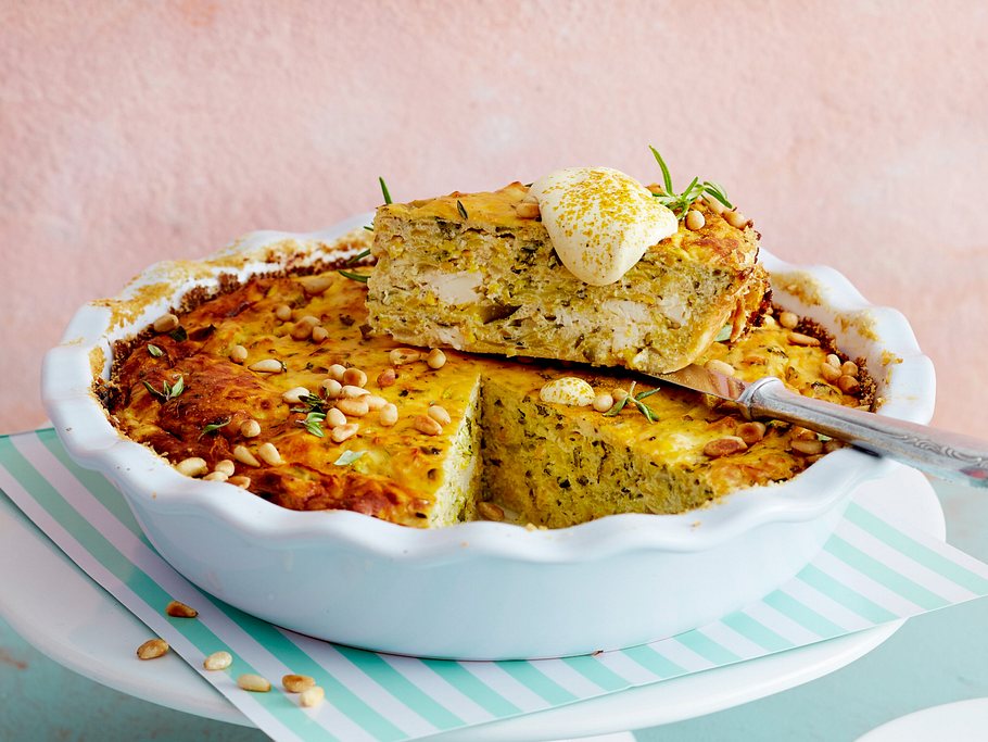 Ein Stück Zucchini-Feta-Kuchen mit Pinienkernen und Kräutern liegt auf einer Kuchenform, darauf ein Klecks Creme.