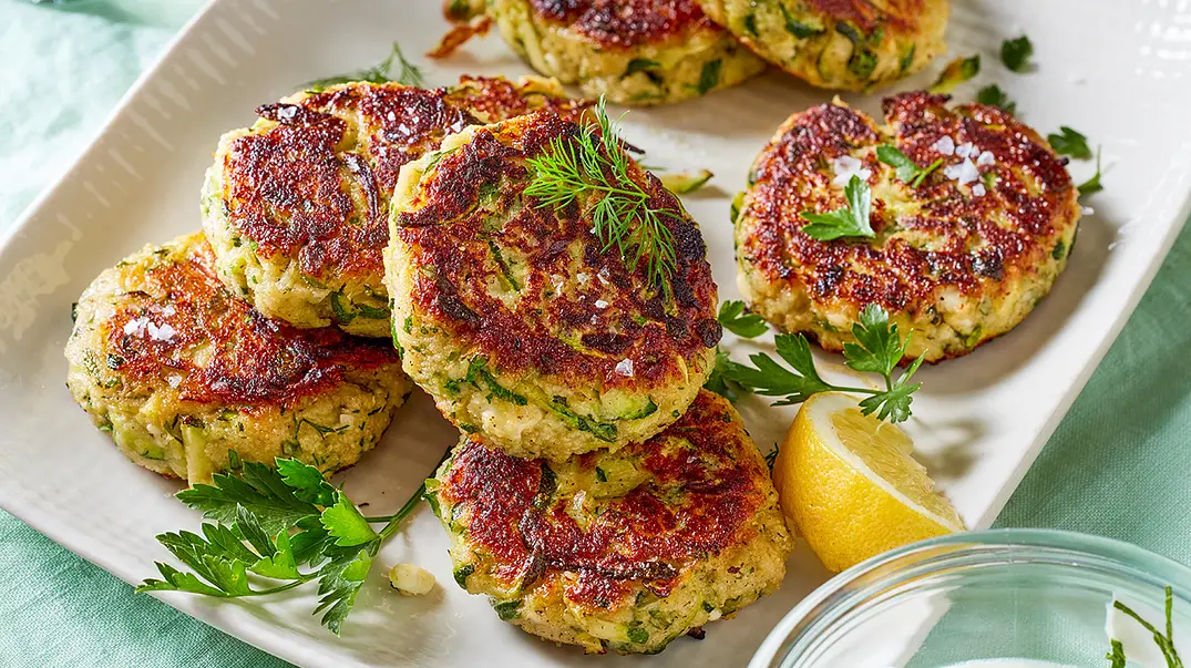 Knusprig gebraten Zucchini-Frikadellen liegen auf einer Servierplatte. Serviert werden sie mit frischen Kräutern, grobem Meersalz und Zitrone. - Foto: LECKER @ Bauer Media Group