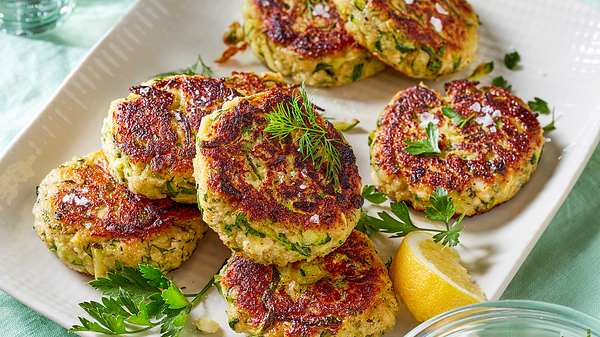 Zucchini-Frikadellen auf einem weißen, rechteckigen Teller mit Zitronenspalte und frischen Kräutern - Foto: House of Food / Bauer Food Experts KG
