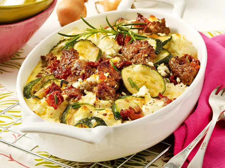 Zucchini-Hackfleisch-Auflauf mit Feta Rezept Der Zucchini-Hackfleisch-Auflauf mit Feta und Reis befindet sich in einer weißen Auflaufform und ist mit einem Zweig Rosmarin garniert.
