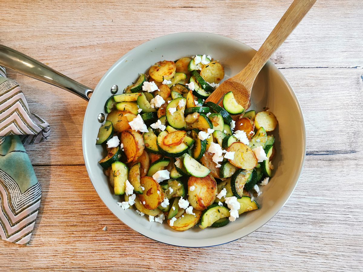 Zucchini-Kartoffel-Pfanne mit Feta bestreut auf einem Holztisch serviert.