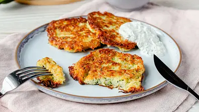 Drei goldbraun gebackene Zucchini-Kartoffel-Puffer neben einem Kräuterdip auf einem Teller - Foto: ShowHeroes für LECKER @ Bauer Media Group