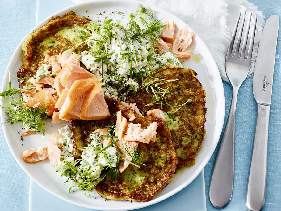 Knusprige Zucchini-Puffer auf einem weißen Teller, serviert mit frischer Kräutercreme und saftigem Stremellachs, garniert mit Dill und Kresse; daneben liegt ein Besteckset auf einer weißen Stoffserviette.