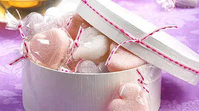 Rosa und weiße Zuckerherzen einzeln in Klarsichtfolie verpackt, dekorativ in einer runden, weißen Geschenkbox auf einem lilafarbenen Tuch arrangiert. - Foto: LECKER @ Bauer Media Group
