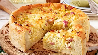 Ein runder und klassischer Zwiebelkuchen mit Speck steht angeschnitten auf einem Holzbrett. - Foto: LECKER @ Bauer Media Group
