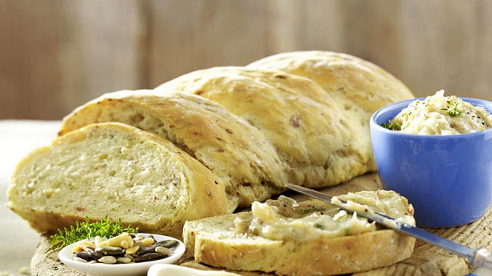 Frisch aufgeschnittenes Zwiebelbrot auf einem rustikalen Holzbrett, serviert mit Aufstrich, gerösteten Kernen und frischen Kräutern. - Foto: House of Food / Bauer Food Experts KG 